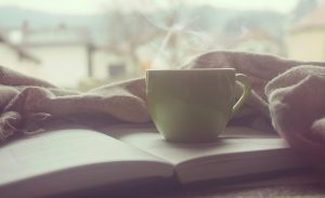 A cup of steamy coffee sitting on an open page of a book with soft focus on a window in the background.