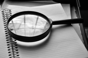 Black and white photo of a magnifying glass resting on a notebook page. Reflection from a nearby window appears on the glass.